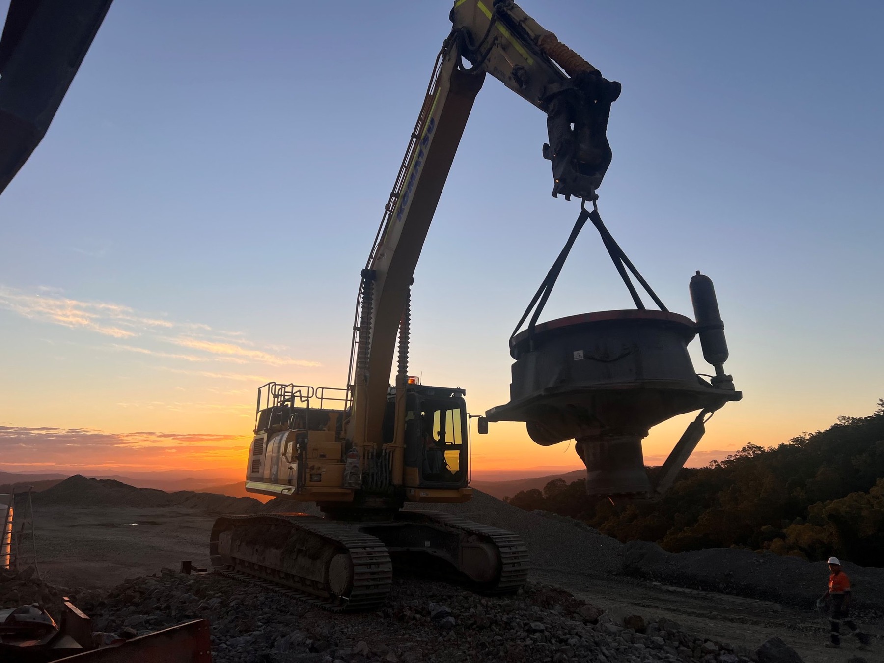 Komatsu excavator lifting a cone crusher head at sunset on a quarry site
